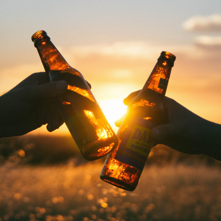 Zwei Hände stoßen mit Bierflaschen gegen den Hintergrund eines Sonnenuntergangs an.