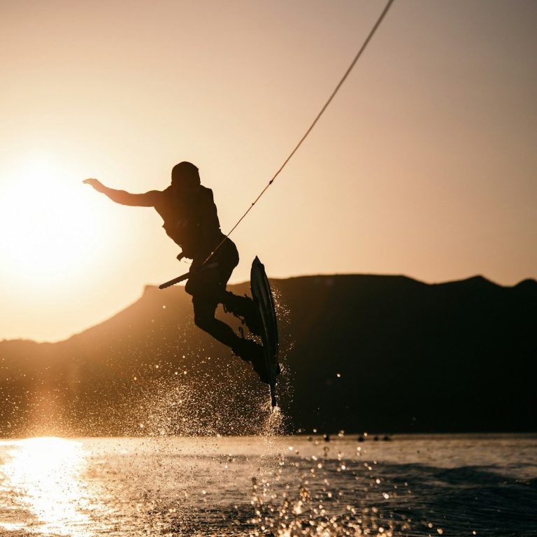 Wakeboarder springt über das Wasser vor einer sonnigen Berglandschaft.