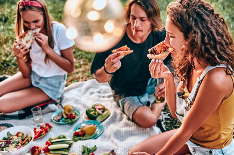 Midsommar Special Drei Personen genießen ein Picknick mit verschiedenen Speisen auf einer Decke im Freien.