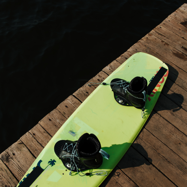 Gelbes Wakeboard mit schwarzem Zubehör auf einem Holzsteg über Wasser.