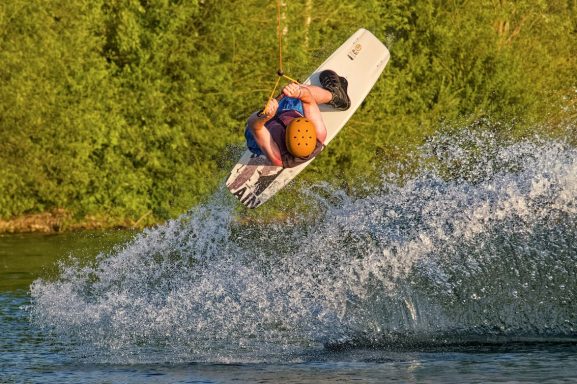 Surfer springt über das Wasser und erzeugt spritzende Wellen im Hintergrund von grünem Ufer.