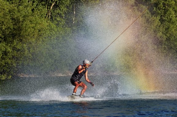 Wasserskifahrer springt über Wellen und ist von einem Regenbogen umgeben.