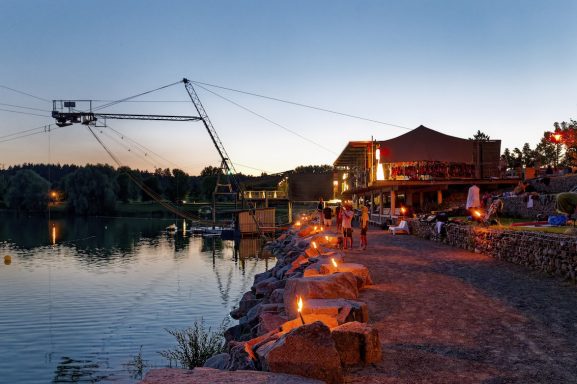 Abendliche Aussicht auf einen See mit Abenteuerpark und illuminierten Uferbereichen.