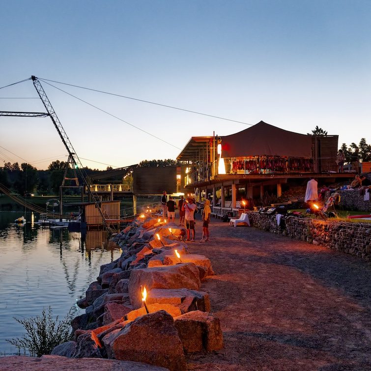 Abendliche Szenerie am See mit Menschen, Fackeln und einem Wassersport-Zentrum im Hintergrund.