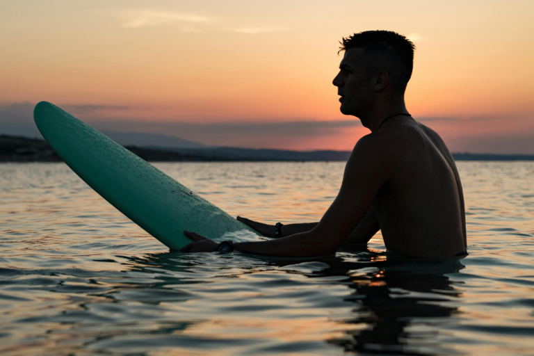 Happy Hour Surfer sitzt im Wasser bei Sonnenuntergang, hält ein Surfbrett.