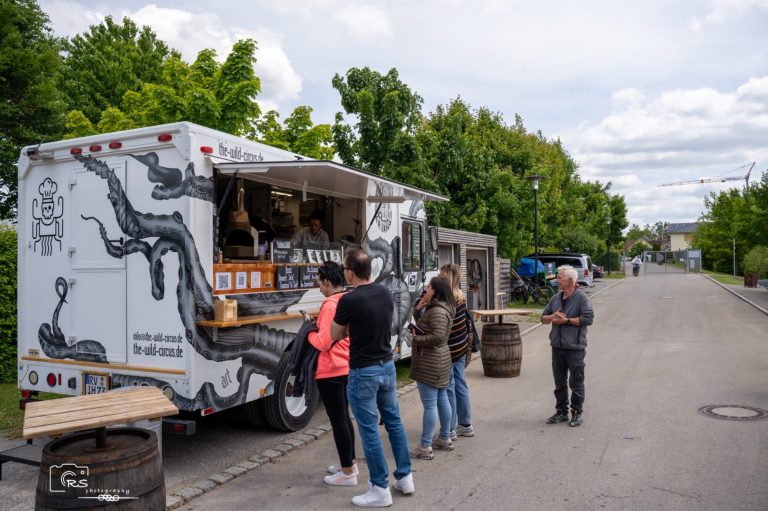 Kostümwettbewerb Ein Foodtruck mit Menschen, die anstehen, um Essen zu bestellen, umgeben von Bäumen.