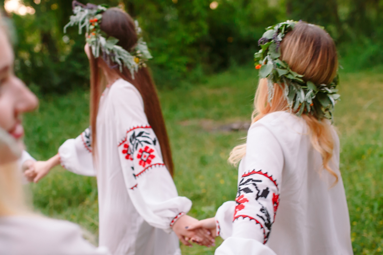 Midsommar Special Zwei Frauen in traditionellen Blusen mit Blumengirlanden, die Händchen halten.